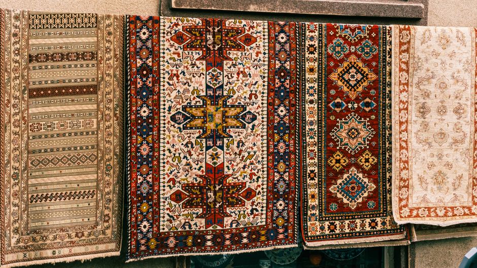 Three vibrantly colored woven rugs with intricate geometric and traditional patterns in red, blue, orange, and white, hung on a wooden exterior wall under an overhang. The rugs hang vertically, reveal