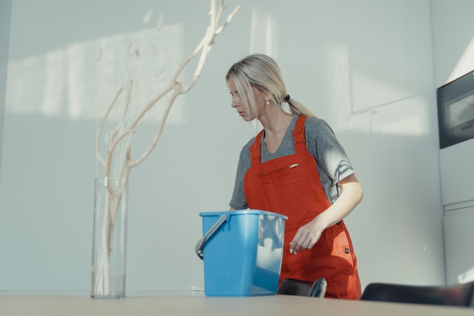 A woman wearing a grey t-shirt and red apron is standing in a well-lit kitchen, heading towards a table with a blue cleaning bucket to perform surface cleaning or deep cleaning as part of a routine fo