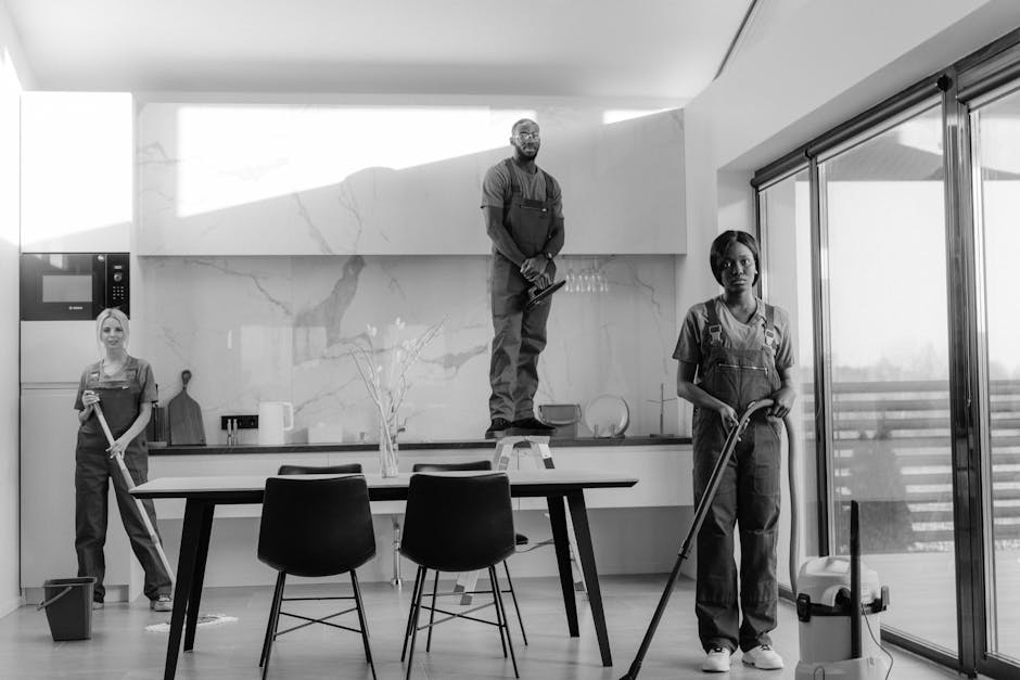 A black-and-white photograph of three professional cleaning workers inside a modern, bright kitchen, actively engaged in surface cleaning and sanitisation. The room features a large white marble backs