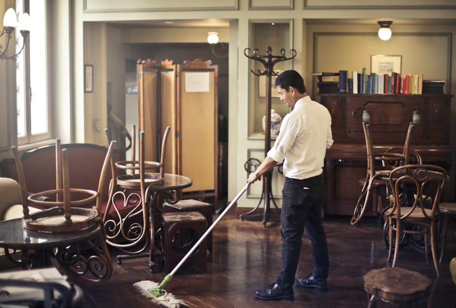 A black-and-white photograph of three professional cleaning workers inside a modern, bright kitchen, actively engaged in surface cleaning and sanitisation. The room features a large white marble backs
